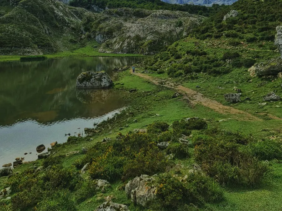 A still mountain lake below rocky peaks, with a footpath running along the shore.